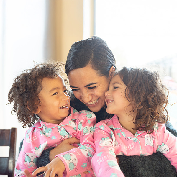 A smiling woman hugging two young children wearing pink patterned pyjamas in a brightly lit indoor setting.