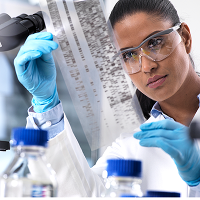 A scientist wearing safety glasses and blue gloves examines a genetic test sequence film in a laboratory setting.