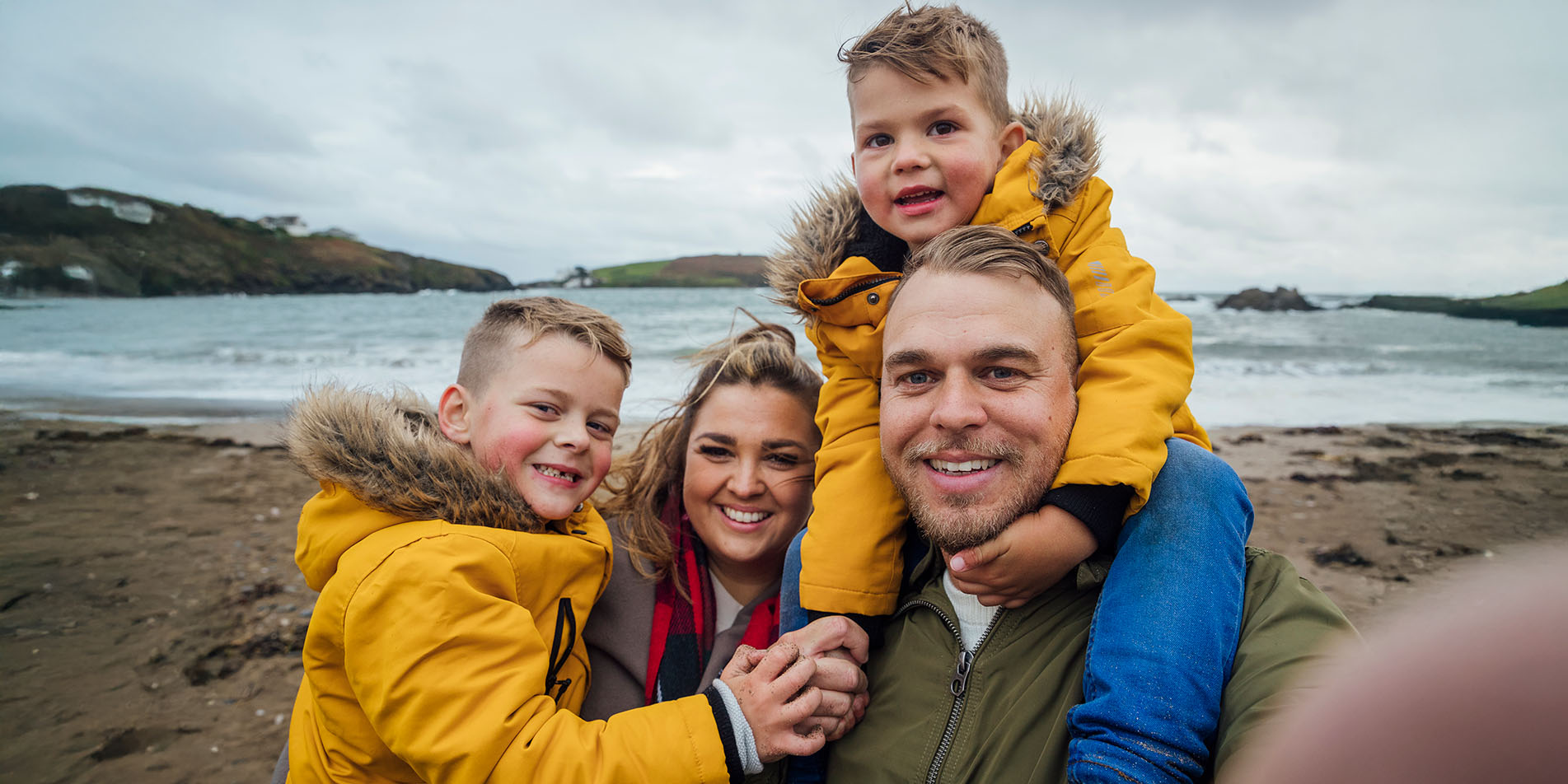 A smiling family of four, including two young children in yellow jackets, posing for a selfie on a sandy beach.